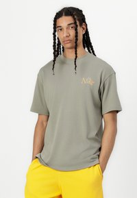 T-shirt en coton vert olive avec un col rond et des manches courtes ; présente un petit logo Nike orange sur la poitrine gauche ; porté avec un short jaune.