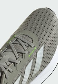Scarpa sportiva grigia con tomaia in rete, dettagli riflettenti bianchi, superficie testurizzata e cuciture verde neon sui lacci.