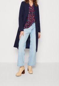 Marineblå lang frakke over en bluse med blomsterprint, lyseblå vidde jeans og beige støvler med blokhæl, der fremhæver teksturer og farver.