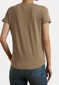 Camiseta marrón de manga corta con cuello redondo y puños enrollados; textura suave y corte relajado, vista desde atrás con jeans de mezclilla.