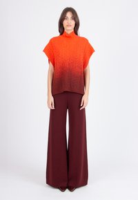 Mujer con suéter de cuello alto de manga corta tejido en degradado de naranja a marrón oscuro y pantalones de pierna ancha color marrón oscuro.