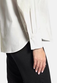 White long-sleeve shirt with a buttoned cuff, featuring a smooth texture and curved hem, worn over black pants with small embellishments.