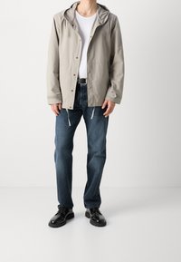 Mężczyzna ubrany w jasnoszary kurtkę z kapturem, białą koszulkę, ciemnoniebieskie dżinsy z czarnym pasekiem i wypolerowane czarne buty wizytowe, stojący na jednolitym tle.