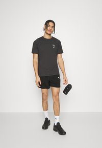 Svart atletisk t-skjorte og shorts med PUMA-logoer, sammen med svarte sko og hvite sokker. Modellen holder en svart vannflaske.
