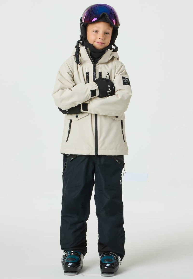 Skijacke in Beige mit schwarzen Akzenten, kombiniert mit schwarzen Skihosen und Stiefeln. Ausgestattet mit Helm und verspiegelter Skibrille.