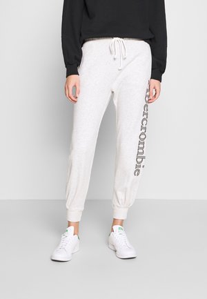 Person iført lysegrå joggingbukser med teksten "Abercrombie" ned ad benet, hvide sneakers og en sort langærmet top mod en ensfarvet baggrund.