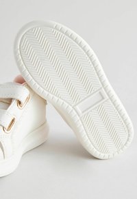 Weiße Sneaker mit einer strukturierten Gummisohle, ausgestattet mit zwei Riemen und goldfarbenen Akzenten. Weiche Innensohle ist an der Öffnung sichtbar.
