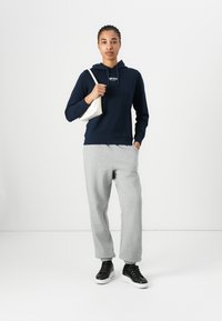 Hoodie marine avec logo sur le devant, pantalon de survêtement gris avec poignets élastiques, et baskets noires. Le modèle porte un petit sac fourre-tout blanc.