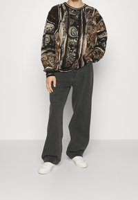 Przewymiarowany sweter w wzory w ziemistych tonach z abstrakcyjnymi motywami; zestawiony z luźnymi ciemnoszarymi dżinsami i białymi trampkami.