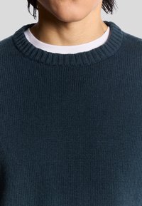 Mörkblå stickad tröja med en ribbad rundad halsringning och en mjuk textur. Lagd över en vit skjorta, som visar upp en enkel, ren design.