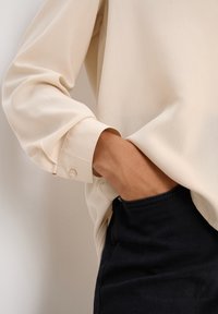 Blouse beige à manches longues avec poignets boutonnés, portée sur un pantalon en denim foncé, mettant en avant une texture lisse et une coupe décontractée.