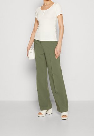Vrouw draagt een wit T-shirt met korte mouwen, olijfgroene wijde broek, witte platformsandalen en houdt een kleine witte handtas vast.