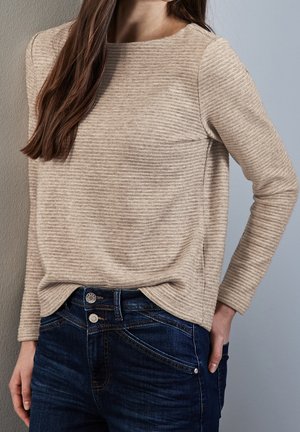 Vrouw draagt een beige, geribbelde trui met lange mouwen, ingestopt in donkerblauwe jeans met dubbele knopen, staand tegen een grijze achtergrond.