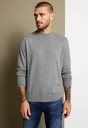 Pull en maille grise avec un col rond et des poignets côtelés. Porté avec un jean bleu, présentant une texture lisse et une coupe ajustée.