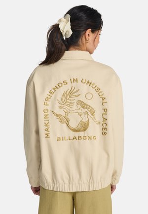 Beżowa kurtka o luźnym kroju, z złotym haftowanym wzorem syreny i napisem na plecach, elastyczne mankiety i talia.