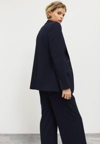 Námořnický oblek na míru, který zahrnuje dlouhý blazer se třemi knoflíky a široké kalhoty. Hladká tkanina s elegantní, strukturovanou siluetou.