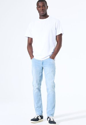 Vaqueros de color azul claro con corte ajustado, combinados con una camiseta blanca lisa. Zapatillas negras y blancas, de pie con las manos en los bolsillos.