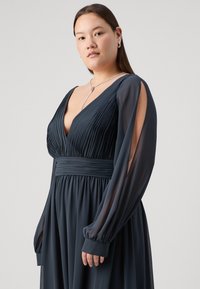 Robe bleu canard foncé avec corsage plissé, décolleté en V profond, manches longues en voile et taille cintrée. Tissu lisse avec jupe fluide. Élégance simple.