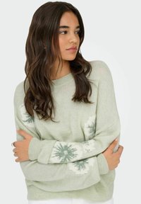 Pull tricoté vert clair avec un col rond, comprenant des motifs de flocons de neige blancs et vert foncé sur les manches et une coupe décontractée.