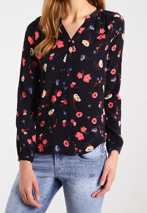 Femme portant un chemisier noir à manches longues avec un imprimé floral rouge, blanc et bleu, associé à un jean déchiré bleu clair.