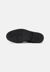 Suela de zapato de goma negra con un patrón de textura. Presenta tacos circulares y rectangulares para proporcionar agarre. Sin marcas ni logotipos visibles.