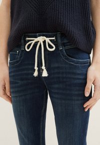 Tmavě modré džíny z denimu s páskem na stažení. Mezi vlastnosti patří přední kapsy, mírně vybledlá textura a kontrastní švy.