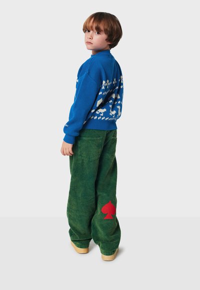 Modrá pletená sveter s bielymi geometrickými vzormi, skombinovaný so zelenými kordovými nohavicami so červeným dizajnom v tvare pika na zadnej strane.