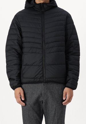 Homme portant une veste noire matelassée à capuche avec une fermeture éclair sur le devant et un pantalon en laine gris, sur un fond uni.
