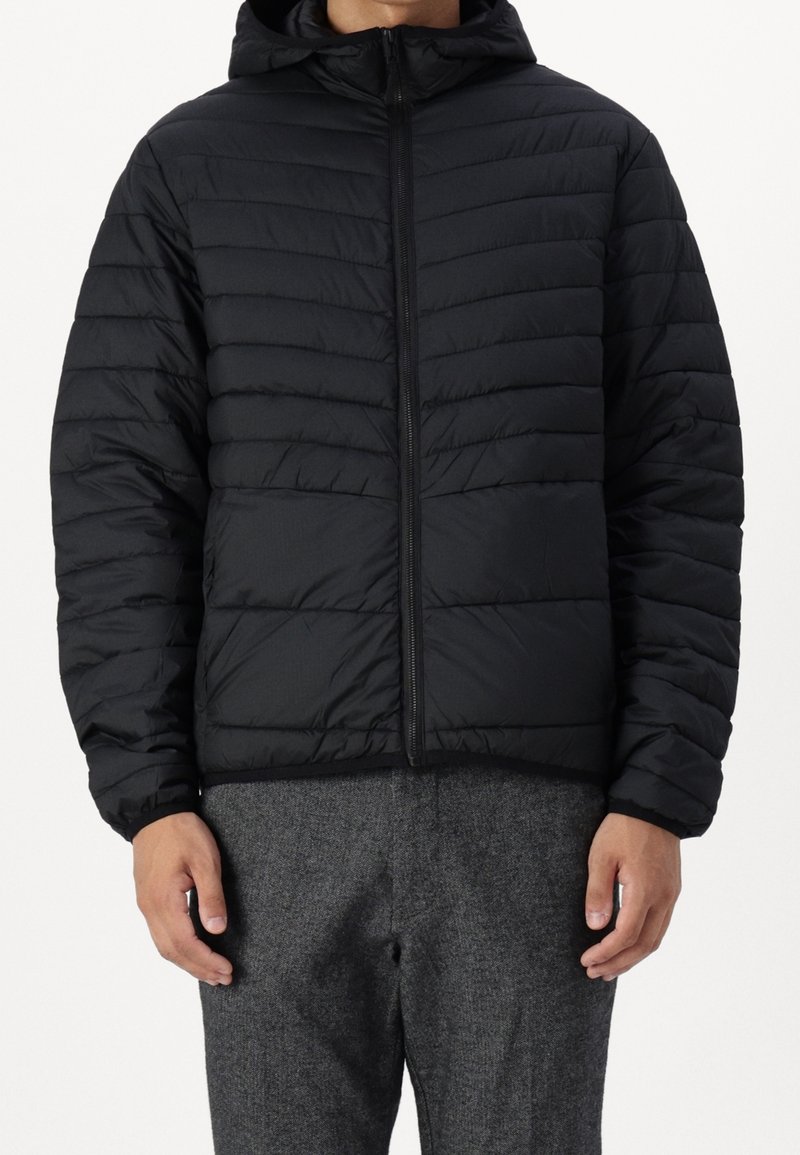 Homme portant une veste noire matelassée à capuche avec une fermeture éclair sur le devant et un pantalon en laine gris, sur un fond uni.
