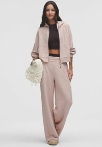 Set de lounge roz deschis, format dintr-o jachetă cu fermoar și buzunare, împreună cu pantaloni largi asortați. Un top scurt maro și o geantă cu șnur de culoare crem completează aspectul.