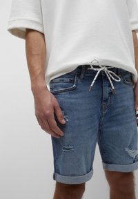 Harmaa ylisuuressa huppari, joka on yhdistetty sinisiin farkkushortseihin, joissa on fringe-reunat ja käännetyt lahkeet, sileä pinta ja nyöriysitys.