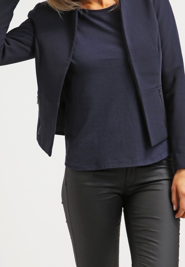 Veste ajustée bleu marine à texture lisse, coupe courte, sans boutons visibles ; dotée de poches latérales zippées. Associée à un tee-shirt bleu marine et un pantalon noir.