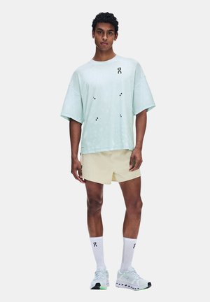 Homme portant un t-shirt oversize bleu clair, un short beige, des chaussettes hautes blanches et des chaussures de course blanches, debout devant un fond uni.