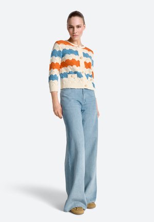 Kobieta ubrana w białą, pomarańczową i niebieską dzianinową marynarkę w wzory z dużymi guzikami, jasnoniebieskie dzwoniaste jeansy i brązowe buty, stojąca.