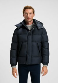 Marinblå puffjacka med dragkedja fram, stor huva och två framfickor. Den har kviltade sektioner och ribbade ärmslut.