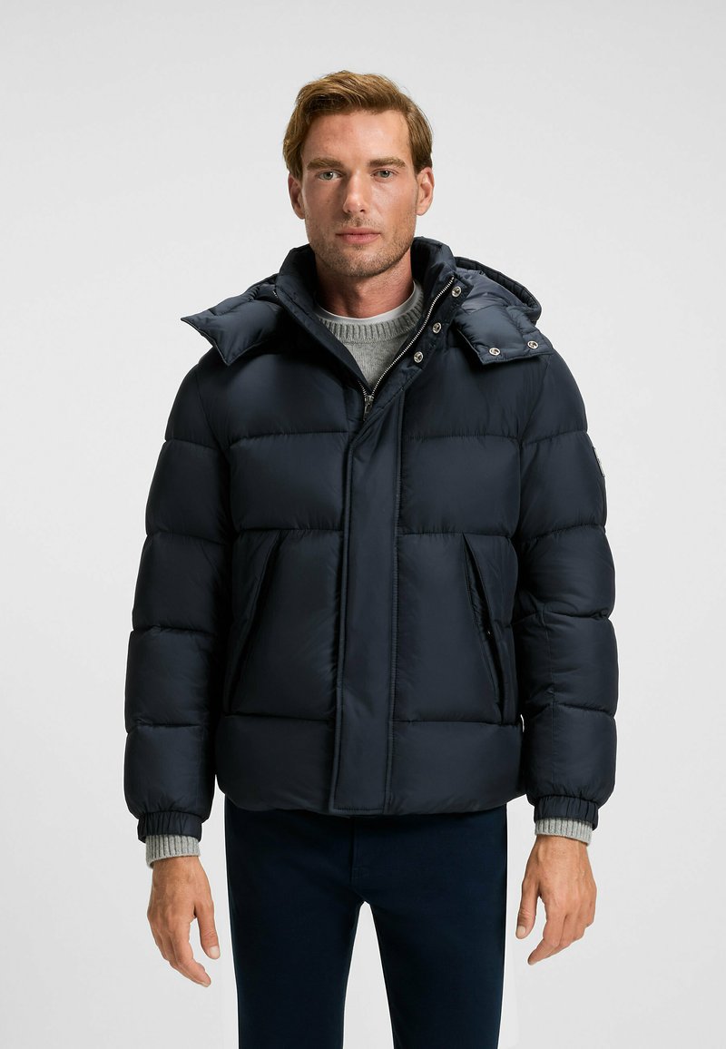 Marinblå puffjacka med dragkedja fram, stor huva och två framfickor. Den har kviltade sektioner och ribbade ärmslut.