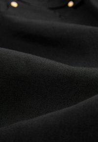 Tissu noir affichant une texture lisse avec un léger éclat. Doté d'un accent en bouton doré sur le col, améliorant son design.