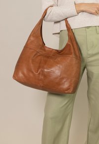 Brun læder hobo taske med en glat tekstur, buet silhuet og en enkelt snoet rem. Stylet med lysegrønne bukser.