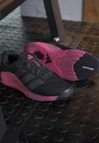 Ζευγάρι μαύρα και ροζ αθλητικά παπούτσια Adidas σε μαύρο υφασμένο πάτωμα γυμναστηρίου με ένα παπούτσι να δείχνει την ροζ σόλα και το λογότυπο Continental.