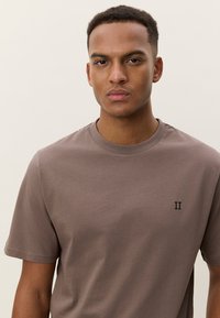 Καφέ βαμβακερό t-shirt με στρογγυλή λαιμόκοψη και κοντά μανίκια. Διαθέτει μικρό σκούρο μπλε λογότυπο "II" στο αριστερό μέρος του στήθους. Λείο ύφασμα.