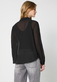 Blusa negra transparente de manga larga con cuello, ajuste holgado y detalle de corte en la espalda. Combinada con jeans gris de cintura alta.