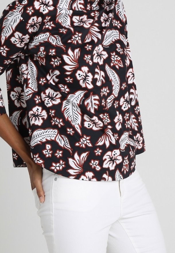 Personne portant une chemise à motifs floraux noir et blanc à manches courtes et un pantalon blanc, main reposant dans la poche.