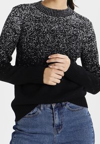 Schwarzer Pullover mit einem gesprenkelten grauen Muster, geripptem Ausschnitt und Bündchen, mit einer lockeren Passform. Wird über Jeans getragen.