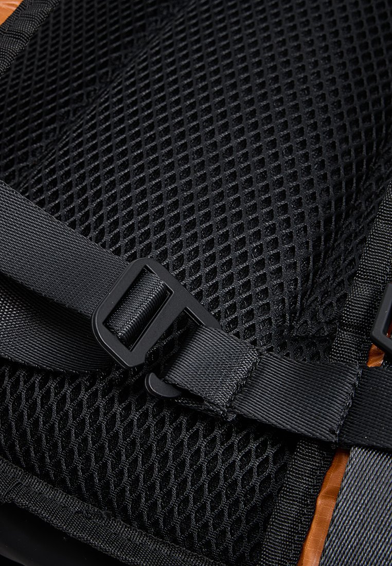 Tessuto a rete nero con motivo a griglia, dotato di spallacci neri regolabili e una fibbia in plastica. È visibile un'elegante accentatura marrone nelle vicinanze.