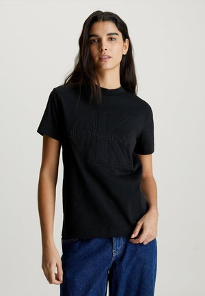 T-shirt nera in cotone con colletto girocollo, caratterizzata da un logo in rilievo nero "Calvin Klein Jeans" sul petto. Indossata con jeans blu.