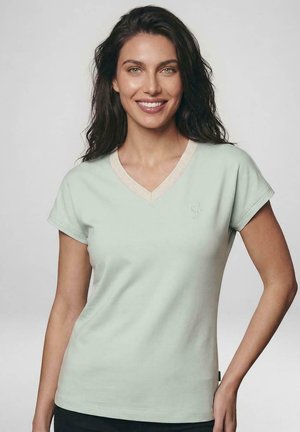 Lachende vrouw met lang donker haar die een lichtgroen V-halshalstshirt met korte mouwen draagt, poseert tegen een effen grijze achtergrond.