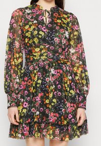 Robe fleurie avec un tissu noir transparent, un motif floral coloré, des manches longues, un décolleté en forme de trou de serrure et un ourlet à volants.