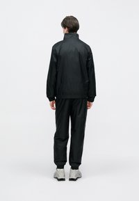 Férfi áll háttal, fekete dzsekit és nadrágot visel, szürke sportcipővel, egyszínű fehér háttér előtt.