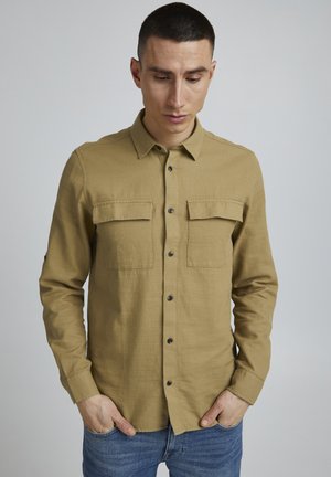 Brown button-up shirt with two front pockets, made of textured fabric, featuring a pointed collar and black buttons. Worn with blue jeans.