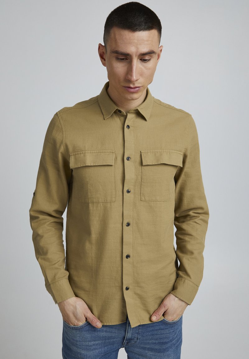 Chemise marron à boutonner avec deux poches avant, en tissu texturé, dotée d'un col pointu et de boutons noirs. Porter avec un jean bleu.